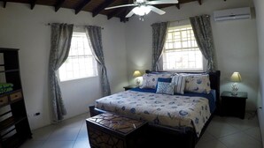 A bedroom with a bed, a ceiling fan, a window with curtains, an air conditioner, and a bedside lamp. - Spacious & Bright 5-bedroom house just a short walk to the beach! (Oistins)