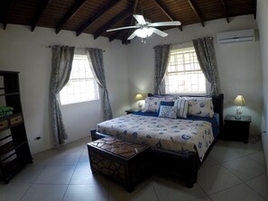 Ein Schlafzimmer mit einem Bett, einem Deckenventilator, einem Fenster mit Vorhängen, einer Klimaanlage und einer Nachttischlampe.