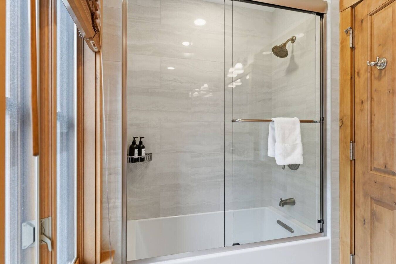 Ducha con cerramiento de vidrio, bañera blanca, armarios de madera y un perchero para toallas.