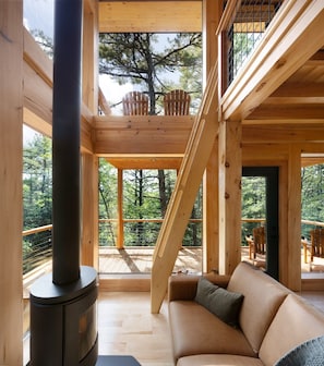 A wooden cabin interior with a sofa, a fireplace, and a balcony with a view of trees. - Forest Views | Private Hot Tub | Alder Treehouse (Ellsworth)