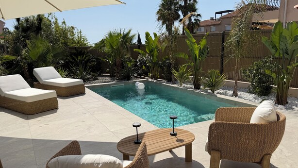 Zona de piscina con mobiliario de mimbre, una mesa y piscina cristalina.