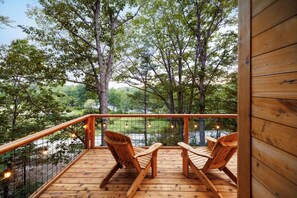 A wooden deck with two wooden chairs, a railing, and a view of trees and a body of water. - River Views | Private Hot Tub | Ash Treehouse (Ellsworth)