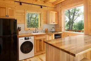 A modern kitchen with wooden cabinetry, a built-in washing machine, and a large window offering a view of trees.