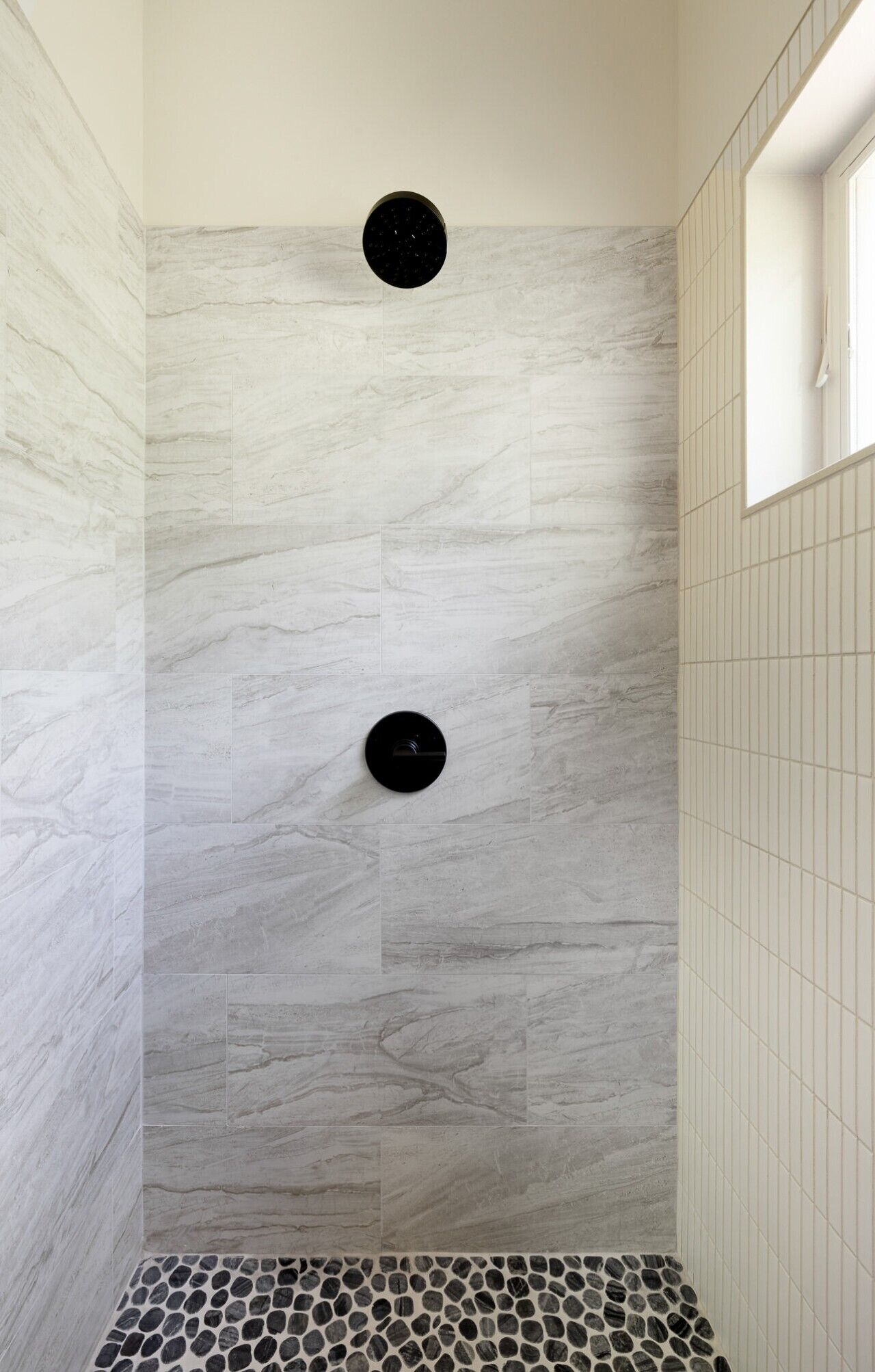 A shower with a tiled wall and floor, featuring a circular drain and a small window.