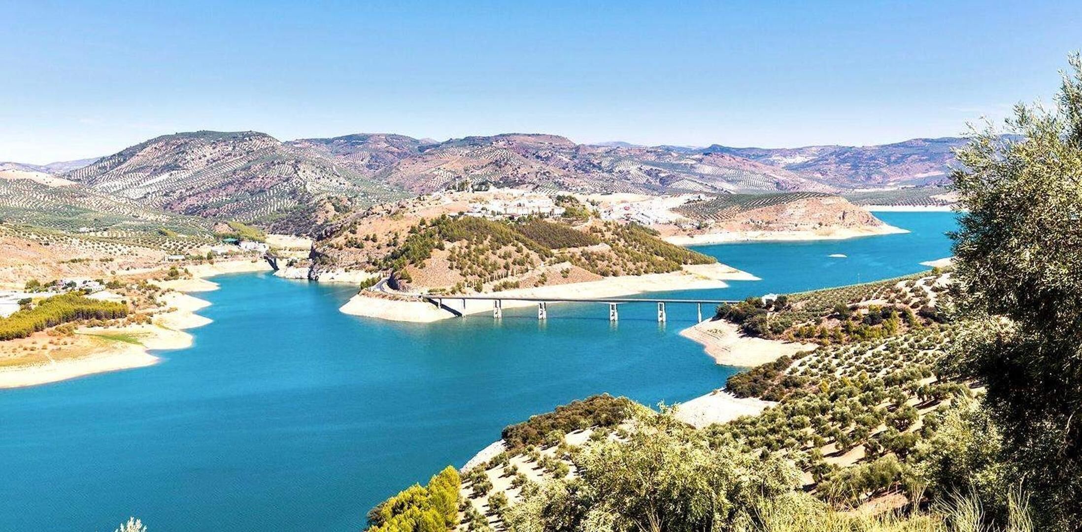 A coastal landscape with a bridge, clear blue water, and hilly terrain.