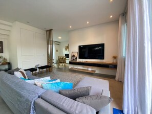 Ein modernes Wohnzimmer mit einem großen Flachbildfernseher, einem grauen Ecksofa und einem Essbereich mit einem hohen Tisch und Stühlen.