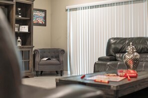 Una sala de estar con un sofá de cuero, un sillón pequeño, una mesa de centro con libros y un jarrón decorativo, y una estantería con cuadros enmarcados y un letrero.