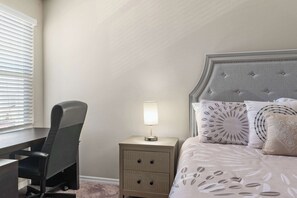 Ein Schlafzimmer mit einem Bett, einem Schreibtisch, einem Stuhl, einem Nachttisch mit Lampe und einem Fenster mit Jalousien.
