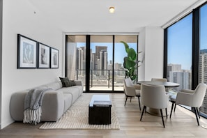 Ein modernes Wohnzimmer mit Sofa, Couchtisch, Essbereich mit Stühlen, großen Fenstern mit Blick auf die Stadt und gerahmten Bildern an der Wand.