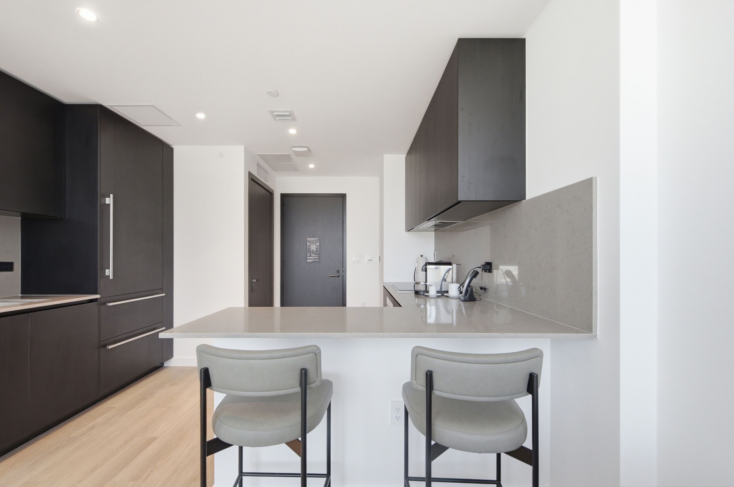 A modern kitchen with dark cabinetry, a central island with two bar stools, and a visible door leading to another room.