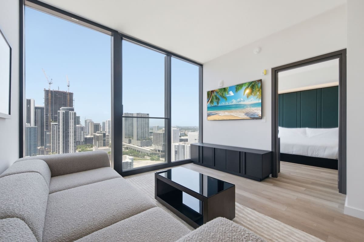 Quarto de hotel moderno com uma janela grande, uma pintura de uma praia, um sofá e uma cama.