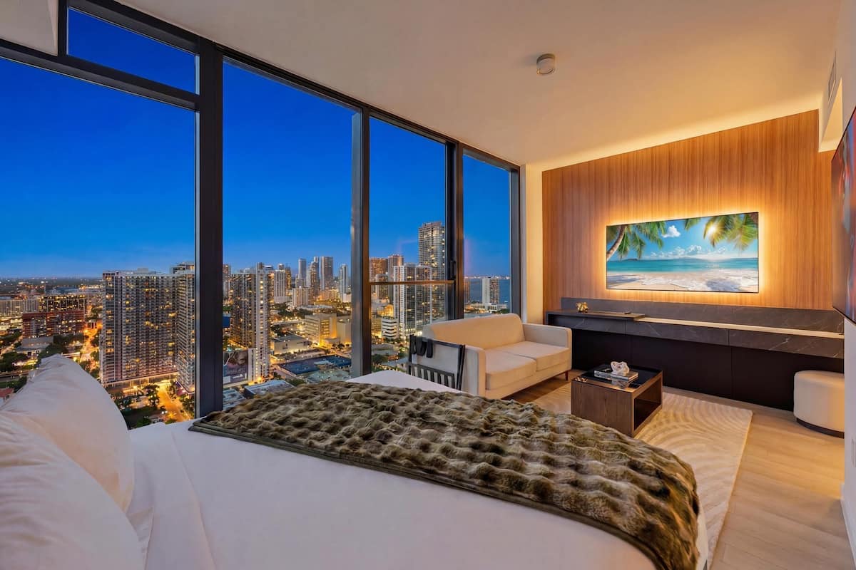 Quarto com uma cama grande, vista para a cidade, televisão na parede e um pequeno espaço para sentar.