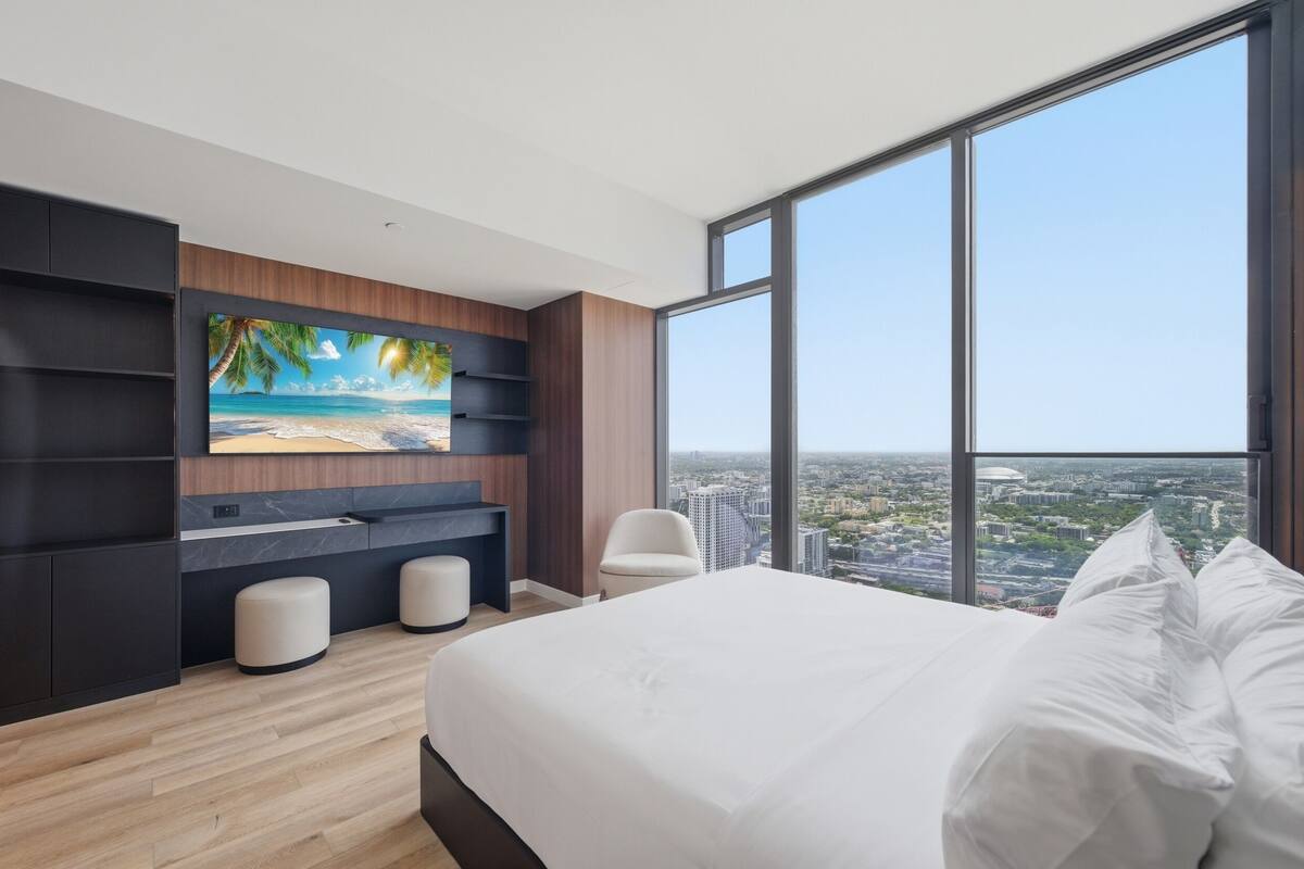 Quarto moderno com uma cama grande, uma TV fixada em uma parede de madeira e vista para a cidade através de grandes janelas.