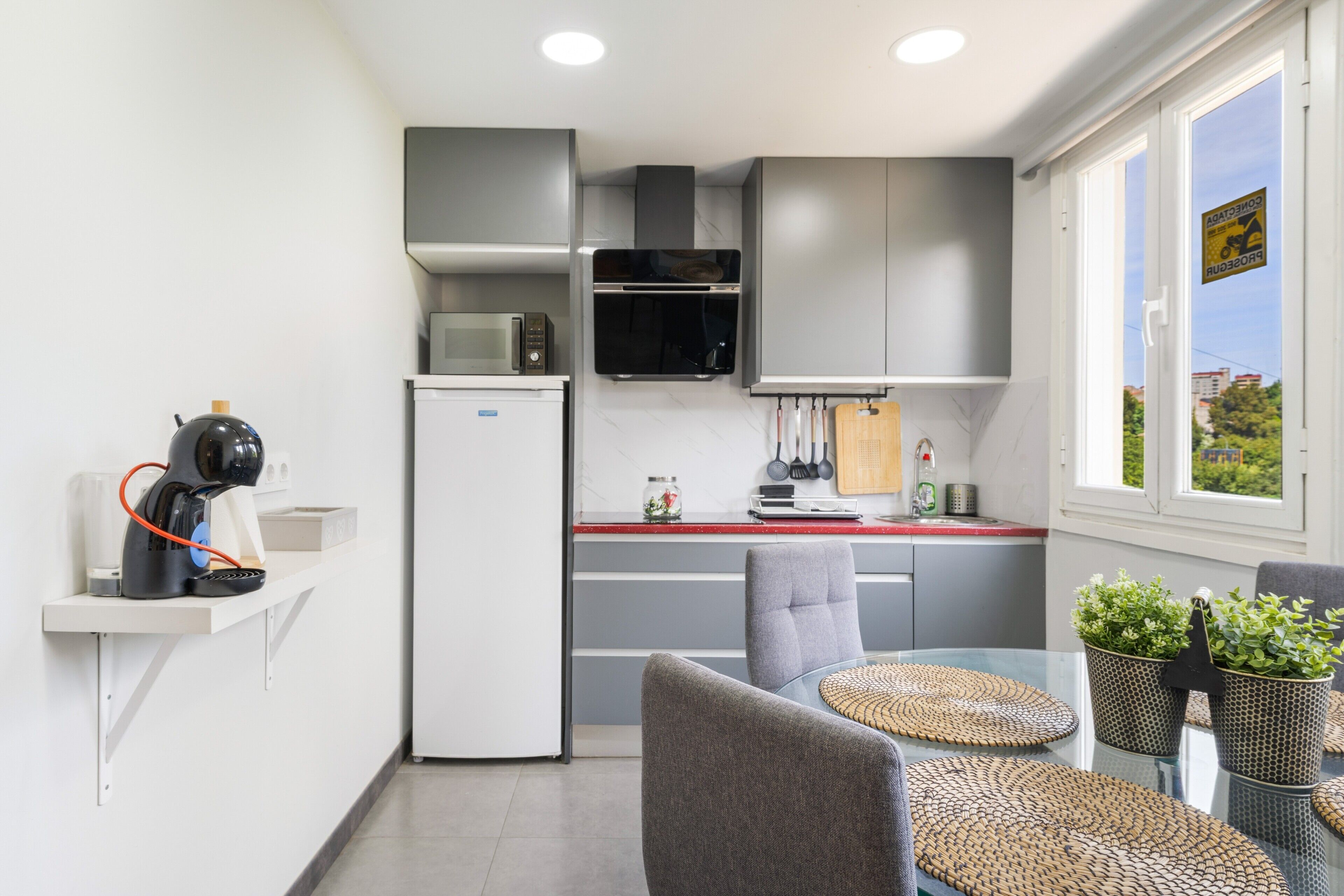 Une cuisine compacte dotée d’appareils électroménagers modernes, un coin repas avec une table et des chaises en verre, et une vue par la fenêtre.