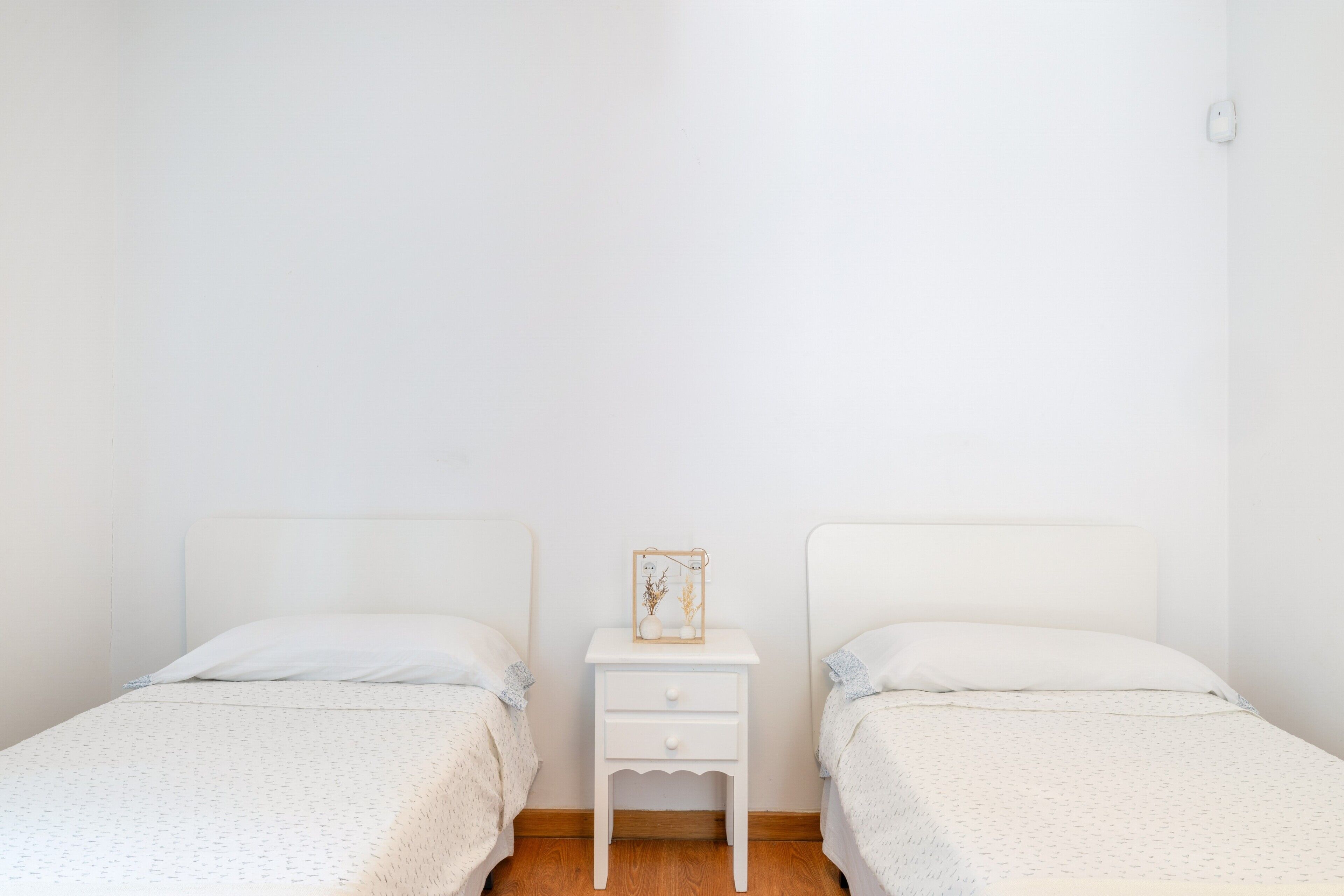 Habitación doble con dos camas individuales, una mesita de noche pequeña con lámpara y una pared blanca lisa.