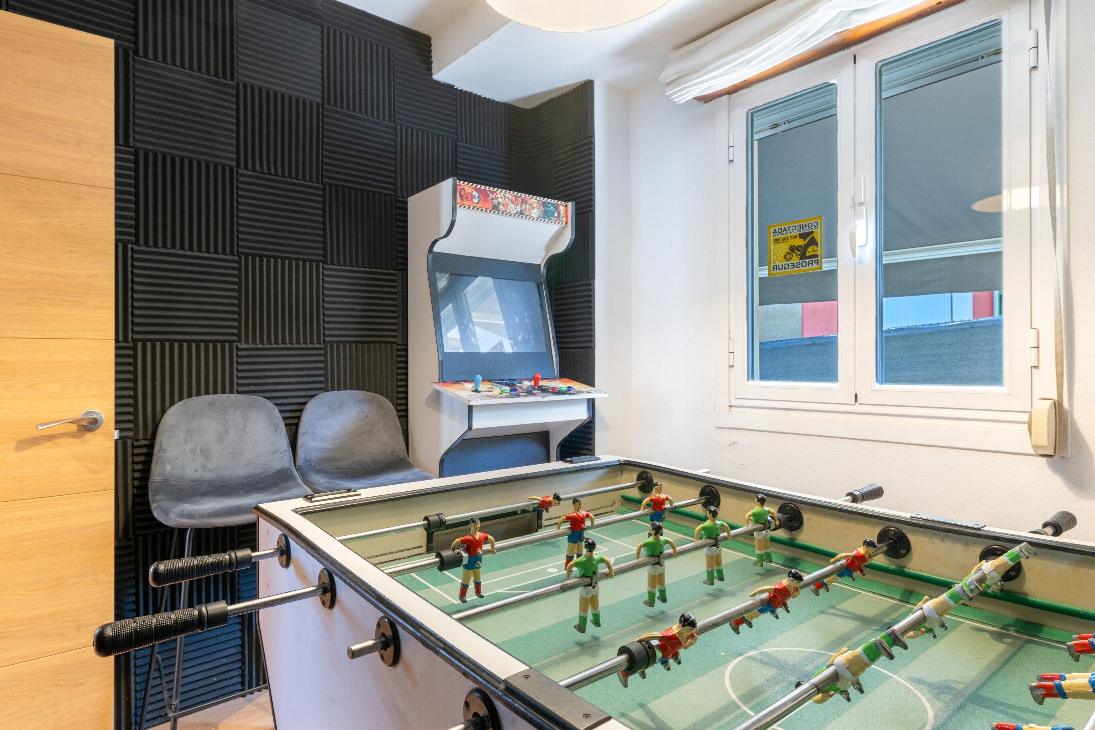 A room with a foosball table, two chairs, a small desk with a computer, and a window with blinds.