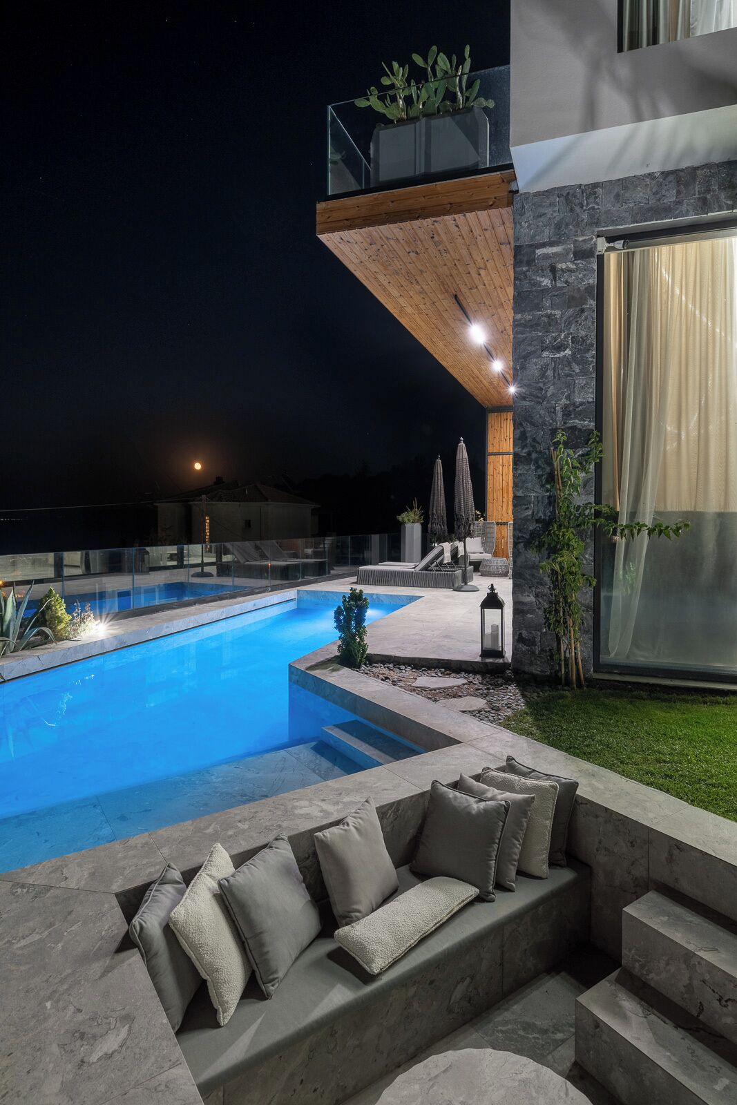 A modern outdoor pool area with a wooden deck, stone walls, and a glass balustrade.