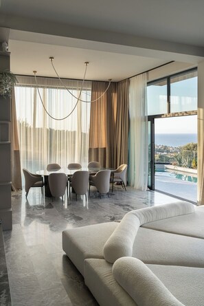 Un salon moderne avec un grand canapé, un coin repas comprenant une table et des chaises, et une vue sur la mer.