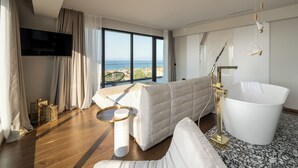 Moderni hotellihuone, jossa on suuri ikkuna, valkoinen kylpyamme, sohva ja litteänäyttöinen televisio.