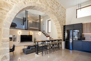 A modern kitchen with a built-in refrigerator, a dining area with a large table and chairs, and a stone archway leading to a living area with a TV and seating. - Villa Methorion Manor - With Private Pool (Saitoures)