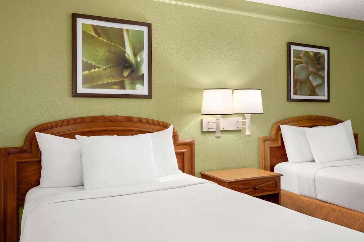 Habitación de hotel con dos camas, una mesita de noche, una lámpara de pared y dos láminas botánicas enmarcadas.