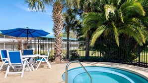 Una piscina con una escalera metálica, rodeada por una cerca, palmeras y sillas azules bajo un paraguas azul.