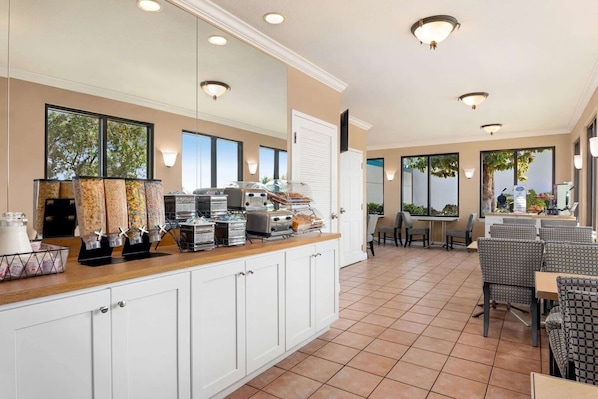 A breakfast bar with cereal dispensers, a coffee machine, and a fruit display. There are chairs and tables in the dining area.