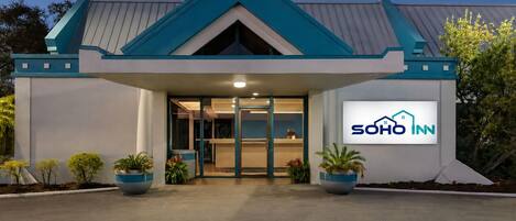 A hotel entrance with a modern design, a visible reception desk, and a logo on the wall.