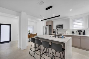 A modern kitchen with a central island, bar stools, a dining area with chairs, and a view of the living room. - Luxury Miami Villa w/ Pool, Jacuzzi, BBQ & Pickleball Court (Miami)