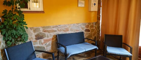 Une pièce avec trois chaises, une table, une fenêtre avec des rideaux, un mur en pierre et un panneau accroché au mur.