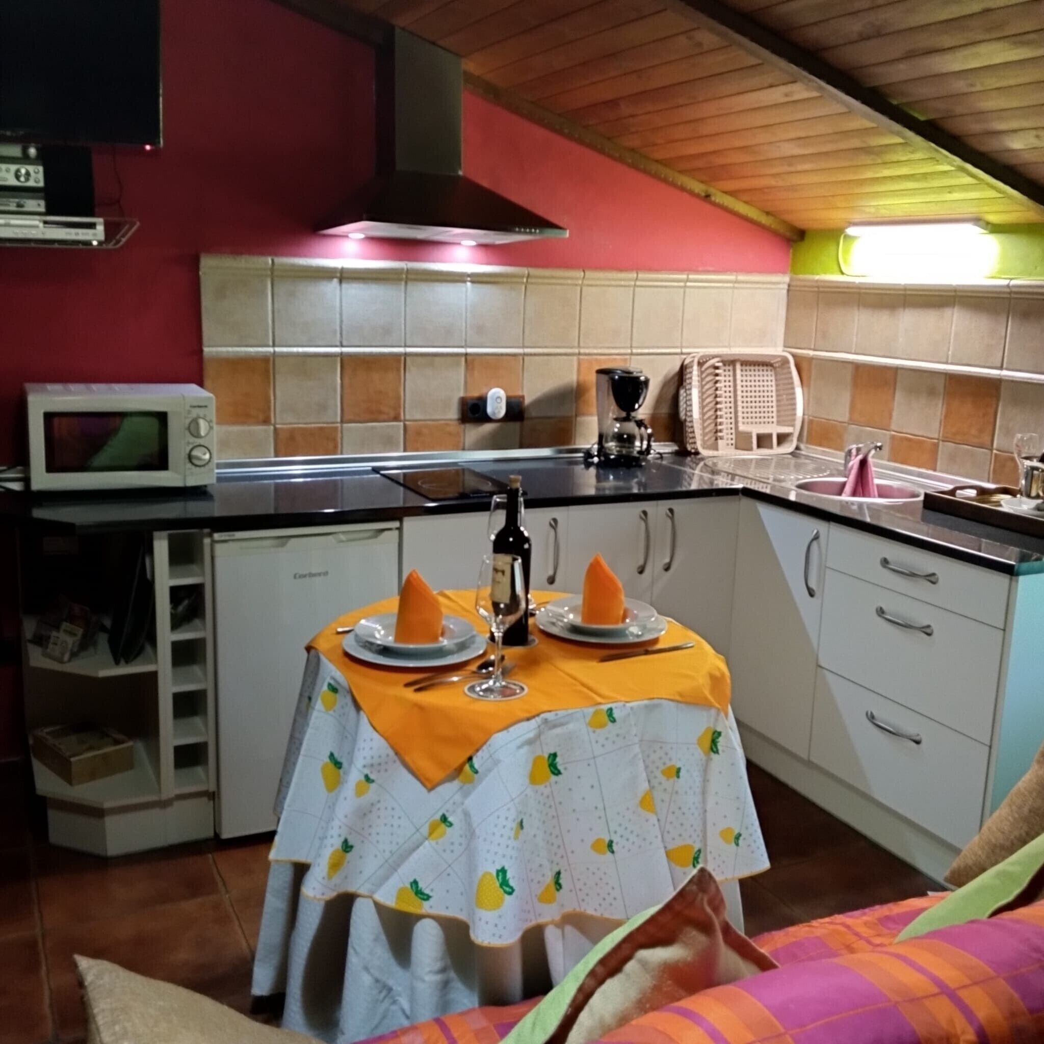 Cocina compacta con microondas, estufa y pileta. Se observa una mesa con mantel y una botella de vino.