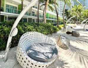 A white, perforated outdoor seating area with a cushion, surrounded by greenery and palm trees, with a modern building in the background. - R&N Azure Staycation (Parañaque)