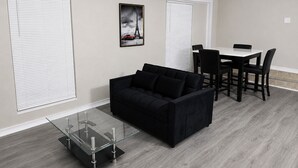 A modern living room with a black sofa, a glass coffee table, a dining table with chairs, and a framed picture of the Eiffel Tower.