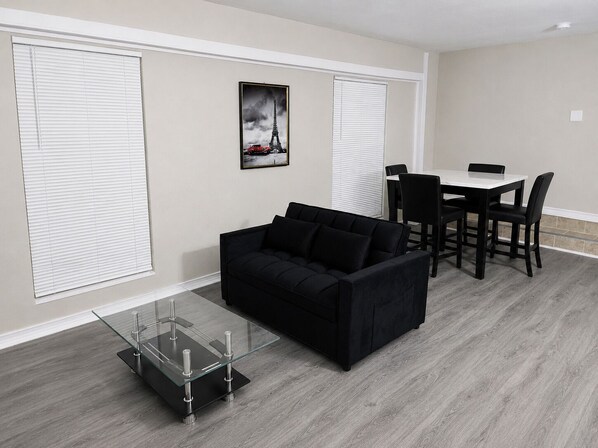 A modern living room with a black sofa, a glass coffee table, a dining table with chairs, and a framed picture of the Eiffel Tower.