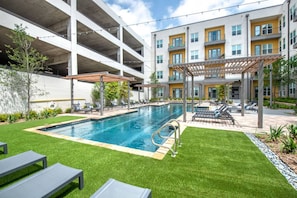 Ein Swimmingpool mit Liegestühlen, einer Pergola und einem mehrstöckigen Gebäude mit Balkonen.