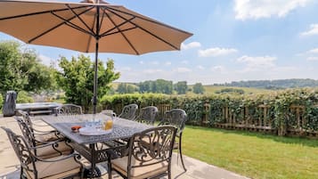 An outdoor patio with a table and chairs, a large umbrella, and a view of a landscaped garden.
