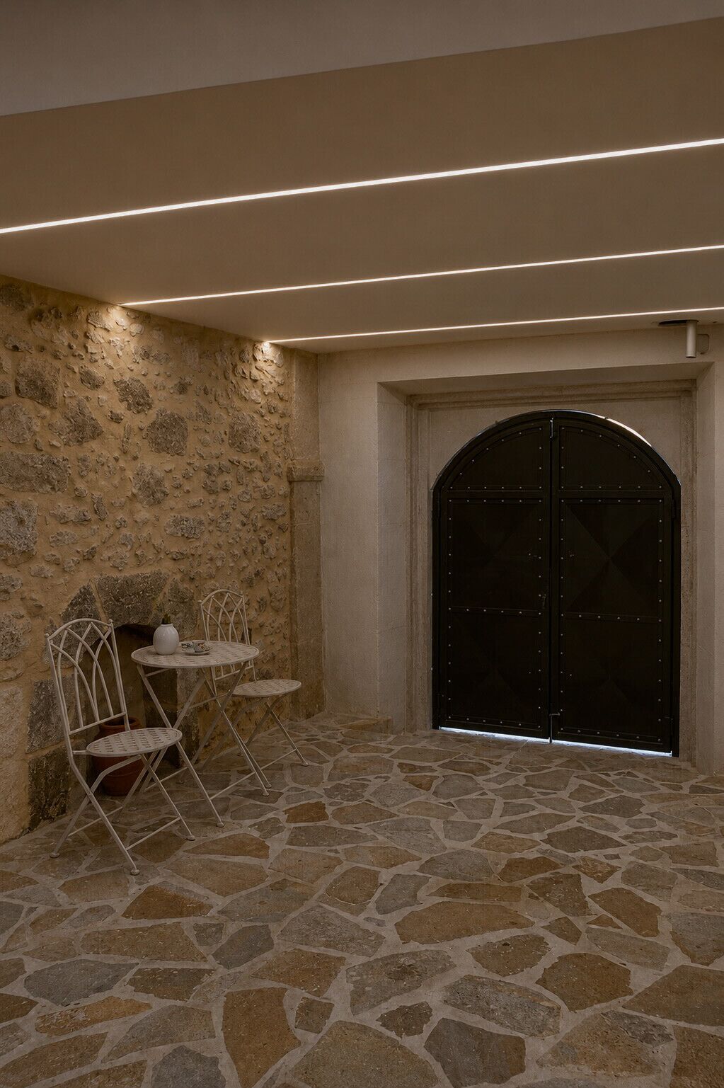 Una habitación con suelo de piedra, una puerta negra, sillas de metal blanco y una pequeña mesa.