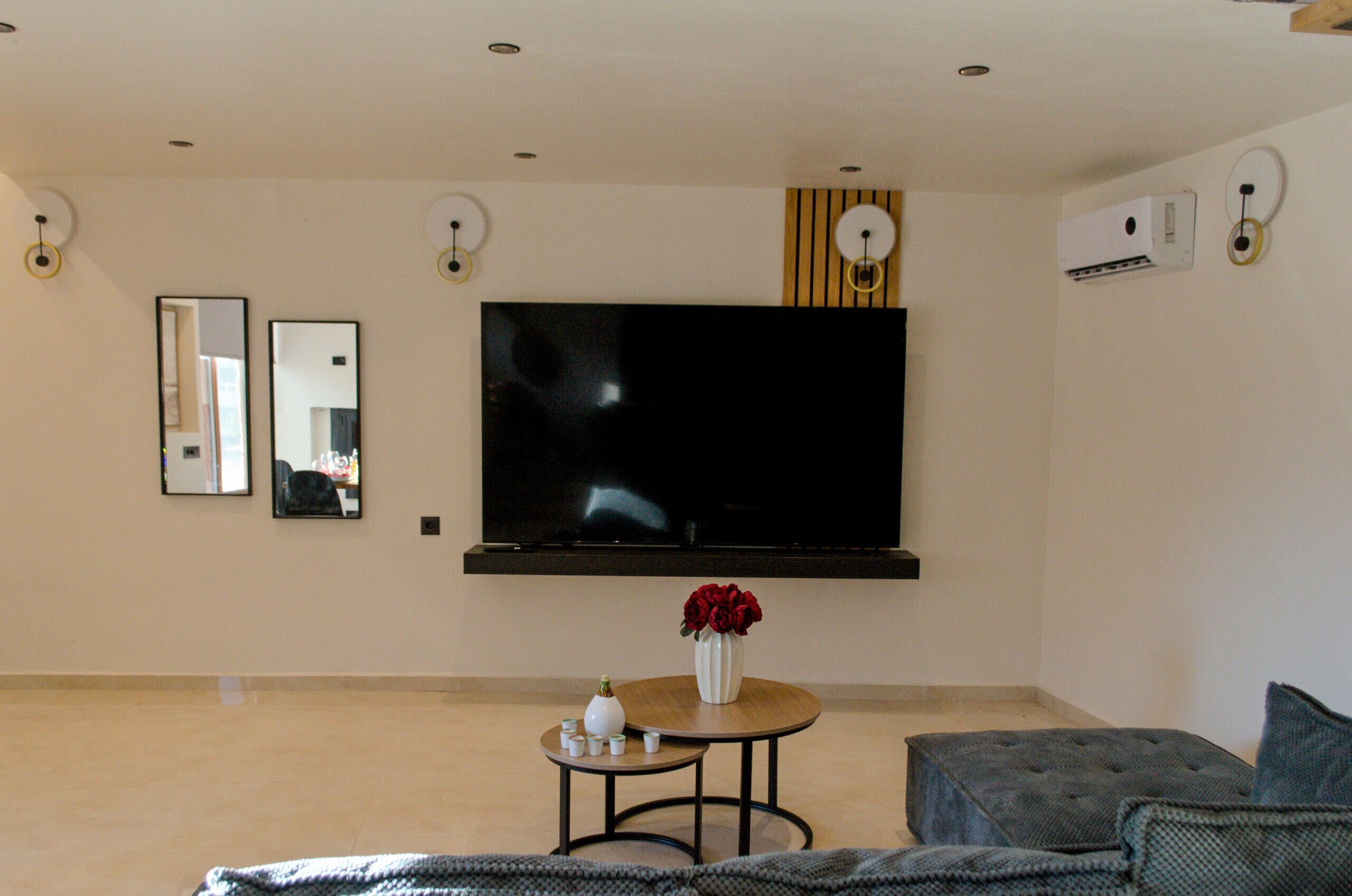 Un salón con televisión de pantalla plana, una mesita redonda con un jarrón de flores rojas, un sofá gris y un espejo grande en la pared.