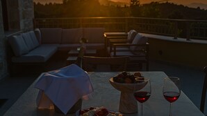 A terrace with a sofa, chairs, a table with a bowl of fruit and two wine glasses, and a sunset in the background. - Villa Methorion Manor - With Private Pool (Rethymno)