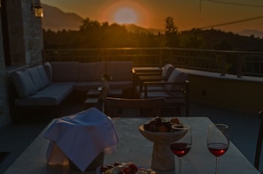 Eine Terrasse mit einer Couch, Stühlen, einem Tisch mit Obstschale und zwei Weingläsern, im Hintergrund ein Sonnenuntergang.