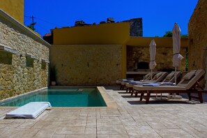Área de piscina com espreguiçadeiras e guarda-sóis, paredes de pedra e um céu azul límpido.