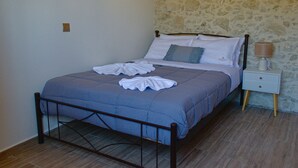 A neatly made bed with a blue bedspread, a white pillow, and a matching pillow on the headboard. There is a bedside table with a lamp and a small drawer. The room has a stone wall and a wooden floor. - Villa Methorion Manor - With Private Pool (Rethymno)