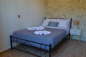 Ein ordentlich bezogenes Bett mit einer blauen Tagesdecke, einem weißen Kopfkissen und einem passenden Kissen an der Kopfteil. Ein Nachttisch mit einer Lampe und einer kleinen Schublade steht daneben. Der Raum hat eine Steinwand und einen Holzboden.