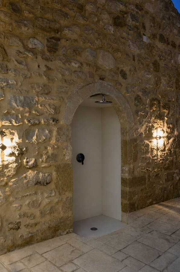 A stone-walled outdoor shower with a simple archway, a metal showerhead, and two wall-mounted lights. - Villa Methorion Manor - With Private Pool (Rethymno)