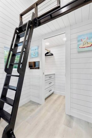 Ein Zimmer mit weißen Wandverkleidungen, einer Holzleiter, einem Gemälde, einem Fernseher und einem Holzkasten.