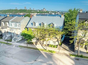 A residential area with a prominent building featuring a steep roof and a large tree in the foreground. - Near the Ferry | L'Appartement des Notaires (Lévis)