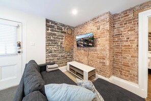 A room with a brick wall, a mounted TV, a sofa, and a door. - Near the Ferry | L'Appartement des Notaires (Lévis)