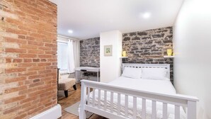 Un dormitorio con cama blanca, pared de ladrillo, escritorio y ventana con persianas.