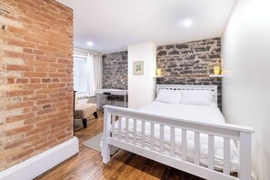 Une chambre avec un lit blanc, un mur en briques, un bureau et une fenêtre avec des stores.