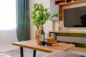 A wooden table with a vase of green leaves, a bottle of wine, and a glass. In the background, there is a television and a shelf with books and decorative items. - 2BR Sunrise Port View Apt Steps from Finikoudes (Larnaka)
